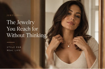 Woman in a cream top fastening a necklace while looking in a mirror.